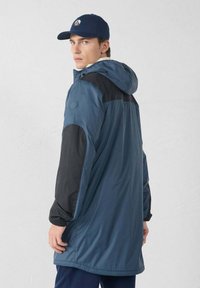 Giacca blu resistente all'acqua con dettagli neri, cappuccio, polsini elastici e una vestibilità rilassata. Presenta una toppa con il logo sulla manica.