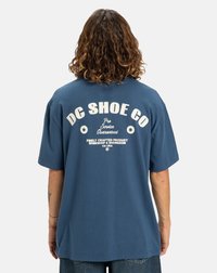 Man med lockigt hår som vänder ryggen mot oss och bär en blå t-shirt från DC Shoe Co med vit text och stjärnor på ryggen, tillsammans med denim jeans.