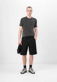 Šedé tričko s krátkym rukávom a čierne cargo šortky. Model drží fotoaparát, na nohách má strieborné tenisky a tmavé ponožky. Jednoduché pozadie.