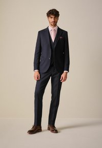 Marineblauwe driescherp pak met een getailleerde pasvorm, bestaande uit een jasje, een gilet en strakke broek, gecombineerd met een lichtroze overhemd en stropdas.