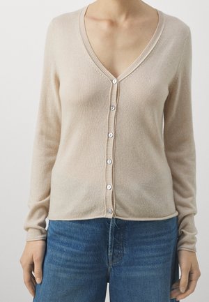 Cárdigan beige con cuello en V y botones, llevado sobre vaqueros vaqueros azules en una persona de pie con los brazos relajados a los lados.