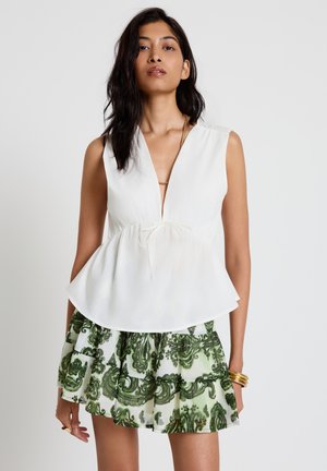 Femme portant un haut péplum blanc sans manches avec un nœud et une jupe verte à imprimé cachemire à volants, debout devant un fond uni.