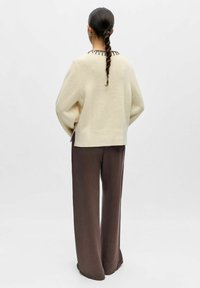 Femme aux longs cheveux tressés portant un pull en maille crème et un pantalon ample marron foncé, debout de dos sur un fond uni.
