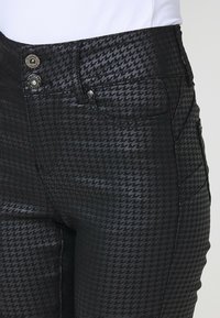 Crne traperice s uzorkom houndstooth, dva gumba za zatvaranje u struku i bočne džepove s glatkom teksturom.