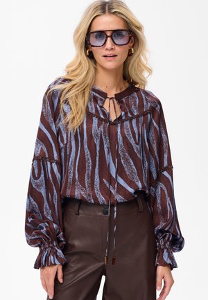 Femme aux longs cheveux blonds portant de grandes lunettes de soleil en écaille de tortue, un blouse à motifs marron et bleu, et un pantalon en cuir marron taille haute.