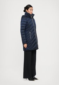 Vrouw staat in profiel, gekleed in een marineblauwe gewatteerde pufferjas met hoge kraag, zwarte brede broek en zwarte schoenen met spitse neuzen en gouden punten.