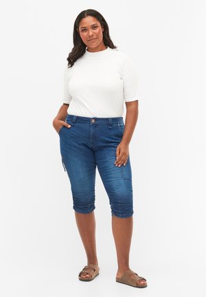 Pantaloni capri in denim blu con una vestibilità aderente, tasche laterali e polsini arricciati, abbinati a una maglietta bianca a maniche corte.