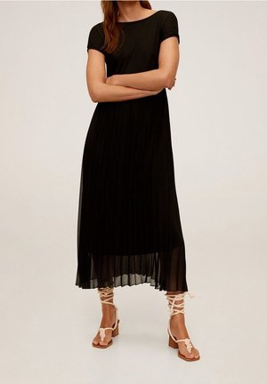 Vrouw draagt een zwart geplooide midi-jurk met korte mouwen en beige sandalen met blokhakken en veter, staand met gekruiste armen.