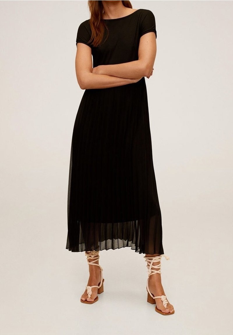 Femme portant une robe midi noire plissée à manches courtes et des sandales à talons blocs beige à lacets, debout les bras croisés.