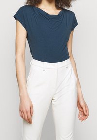 Kvinna klädd i en mörkblå kortärmad topp med draperad halsringning, instoppad i högmidjade vita skräddarsydda byxor, stående mot en enfärgad bakgrund.