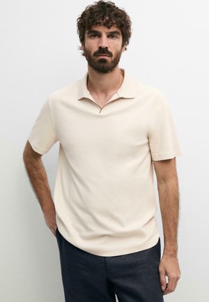 Homme aux cheveux bouclés et à la barbe, portant un polo crème à manches courtes et un pantalon foncé, debout devant un fond clair uni.