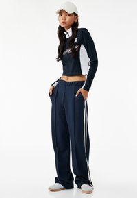 Conjunto de chándal en azul marino y blanco que incluye una chaqueta de cremallera corta con logo y pantalones largos de pierna ancha con franjas laterales, combinado con zapatillas blancas y una gorra.