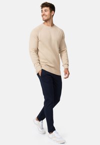 Suéter de punto beige con dobladillo y puños de canalé, combinado con pantalones ajustados azul marino y zapatillas blancas con acentos beige. Conjunto informal en general.