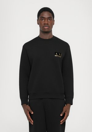 Man standing wearing a black Armani Exchange sweatshirt with small gold logo on left chest against plain light background.