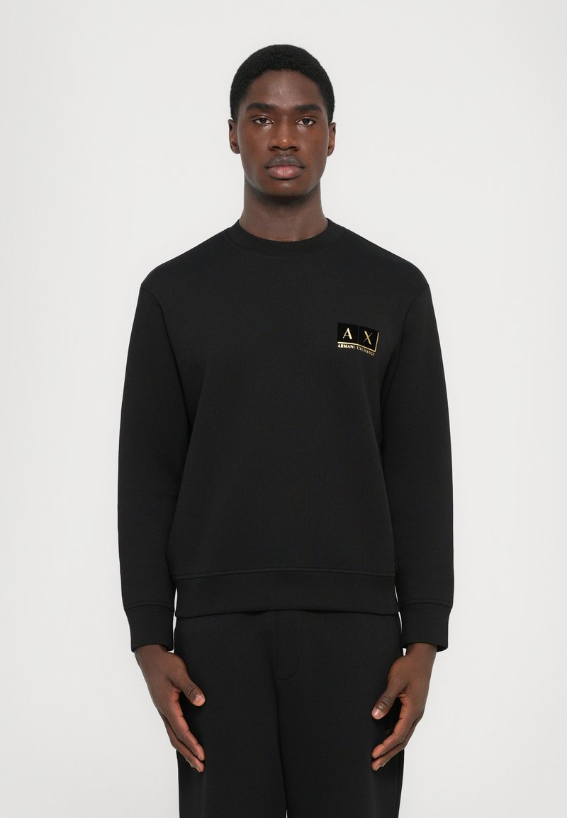 Man standing wearing a black Armani Exchange sweatshirt with small gold logo on left chest against plain light background.