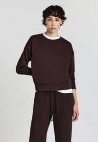Felpa corta marrone realizzata in materiale morbido con scollo rotondo, maniche lunghe e una cintura larga, abbinata a pantaloni coordinati.