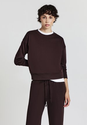 Felpa corta marrone realizzata in materiale morbido con scollo rotondo, maniche lunghe e una cintura larga, abbinata a pantaloni coordinati.