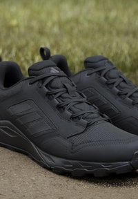 Coppia di scarpe sportive nere con tomaie in mesh e materiale sintetico, caratterizzate da dettagli cuciti e lacci, posizionate su una superficie di cemento con sfondo di erba.