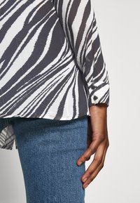 Persona che indossa una blusa a strisce zebrate bianca e nera con polsini abbottonati e jeans in denim blu, con la mano rilassata al proprio fianco.