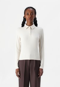 Ribbet langermet polo skjorte i off-white, med knappede krage, strukturert stoff. Kombinert med høytlivede mørkebrune bukser.