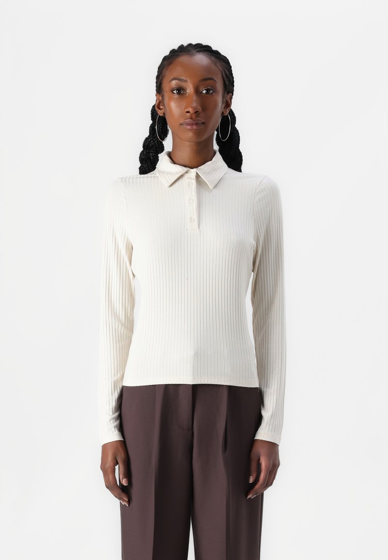 Ribbet langermet polo skjorte i off-white, med knappede krage, strukturert stoff. Kombinert med høytlivede mørkebrune bukser.