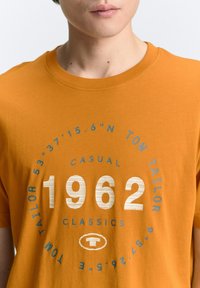 Camiseta de algodón naranja con un diseño impreso en forma redonda que presenta los números "1962" y "TOM TAILOR" en azul y blanco. Estilo casual, con mangas cortas.