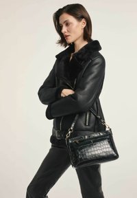 Czarna skórzana kurtka z owczej wełny z zamykanymi kieszeniami; modelka trzyma czarną, teksturowaną torebkę z sztucznej skóry krokodyla z srebrnymi akcesoriami.