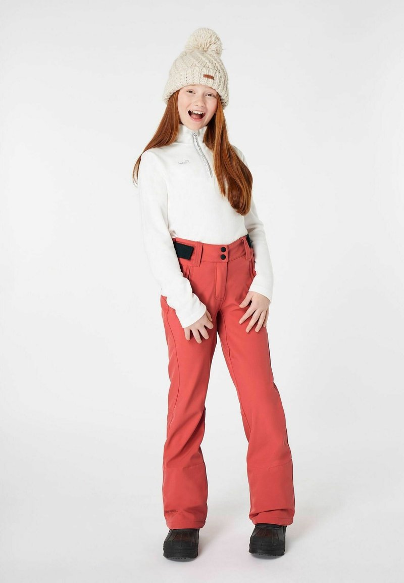 Rote Schneehose mit schwarzen Akzenten, kombiniert mit einem weißen Langarmshirt. Accessoires mit einer cremefarbenen Strickmütze mit Bommel.