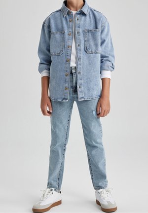 Person iført lyseblå denimjakke med træknapper, hvid skjorte, matchende lyseblå jeans og hvide sneakers med brune såler.