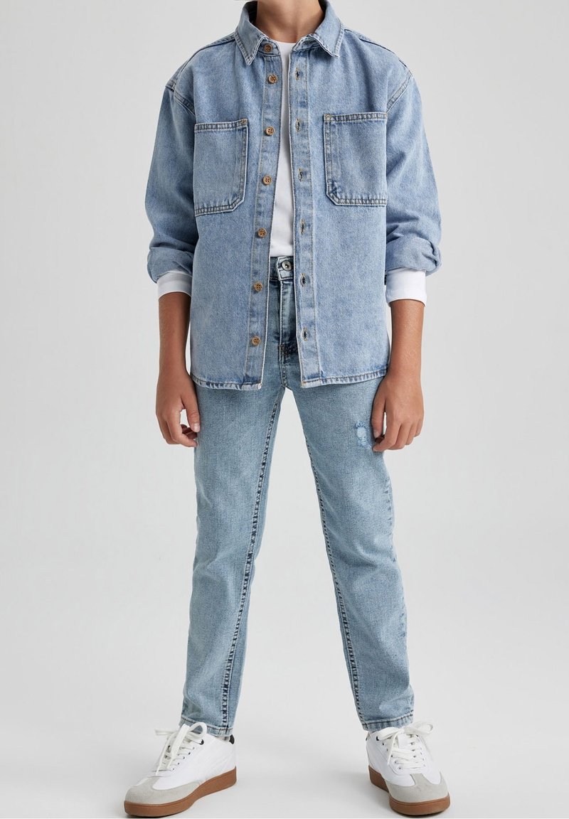 Personne portant une veste en jean bleu clair avec des boutons en bois, une chemise blanche, un jean bleu clair assorti et des baskets blanches avec des semelles marron.