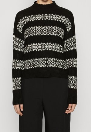 Pull en maille noire avec des rayures horizontales blanches à motif géométrique porté par une personne avec un pantalon noir sur un fond uni.