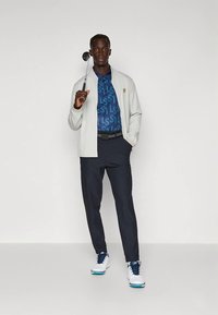 Světlě šedá bunda přes modré polo s vzorem, tmavé kalhoty a bílé golfové boty. Muž drží golfovou hůl. Jednoduchý design a textura.