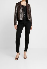 Braune Lederbikerjacke mit strukturierter Oberfläche, ausgestattet mit silbernen Reißverschlüssen und Knöpfen, kombiniert mit schwarzen, enganliegenden Hosen und Riemchenabsätzen.