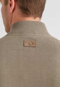 Hellbraun gestrickter Pullover mit einem gerippten Kragen und einem rechteckigen, hellbraunen Lederetikett auf der Rückseite, das geprägte Designdetails aufweist.