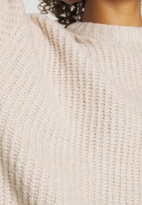 Gros plan d'une personne portant un pull tricoté texturé de couleur crème, mettant en valeur le motif détaillé du tissu et l'encolure.