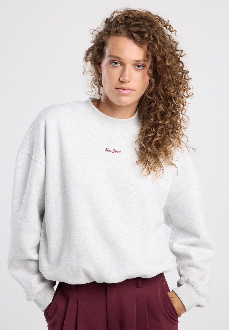 Sweatshirt gris avec des poignets et une taille côtelés. "New York" brodé en rouge sur la poitrine. Modèle associé à un pantalon bordeaux.