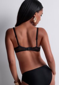 Soutien-gorge en dentelle noire avec des bretelles ajustables, présentant un motif en chevrons et une fermeture à crochets. Associé avec un bas de bikini noir uni.