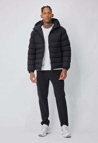 Veste noire matelassée avec capuche, portée sur un t-shirt blanc, associée à un pantalon cargo sombre et des baskets blanches, posant devant un fond clair.