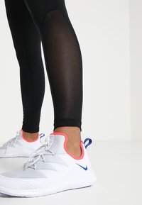 Svarta atletiska leggings med en slät yta och nätpaneler nära knäna, kombinerade med vita sneakers med rosa och blå detaljer.