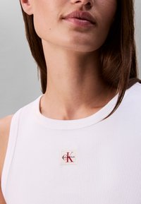 Close-up af en kvinde iført en hvid ribbet tanktop med et lille Calvin Klein logo på brystet.