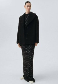 Donna che indossa un cappotto nero oversize su un vestito nero a pois, in piedi con scarpe bianche contro uno sfondo chiaro uniforme.
