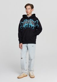 Felpa nera con grafica 'REFLECTION' in blu elettrico, tasca a marsupio frontale, abbinata a jeans di denim chiaro e sneaker beige.