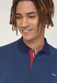 Polo blu navy con tessuto strutturato, presenta un colletto e una patta a righe rosse, e un logo argentato ricamato sul petto.