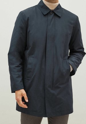 Muž v tmavě modrém zapnutém kabátu přes béžový rolák s jednou rukou v kapse.