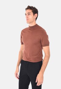 Uomo che indossa un maglione a maniche corte color marrone con colletto alto e pantaloni neri, in piedi e guardando a sinistra contro uno sfondo neutro.