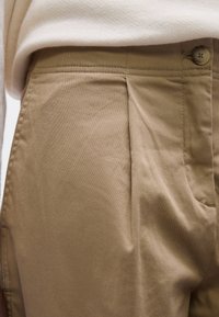 Pantalon beige avec plis sur le devant et fermeture par boutons, associé à un haut beige clair rentré à la taille.