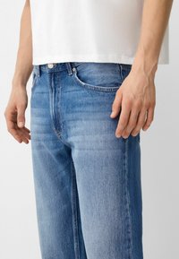 Primer plano de una persona que lleva vaqueros de denim azul claro y una camisa blanca, con las manos relajadas a los lados.