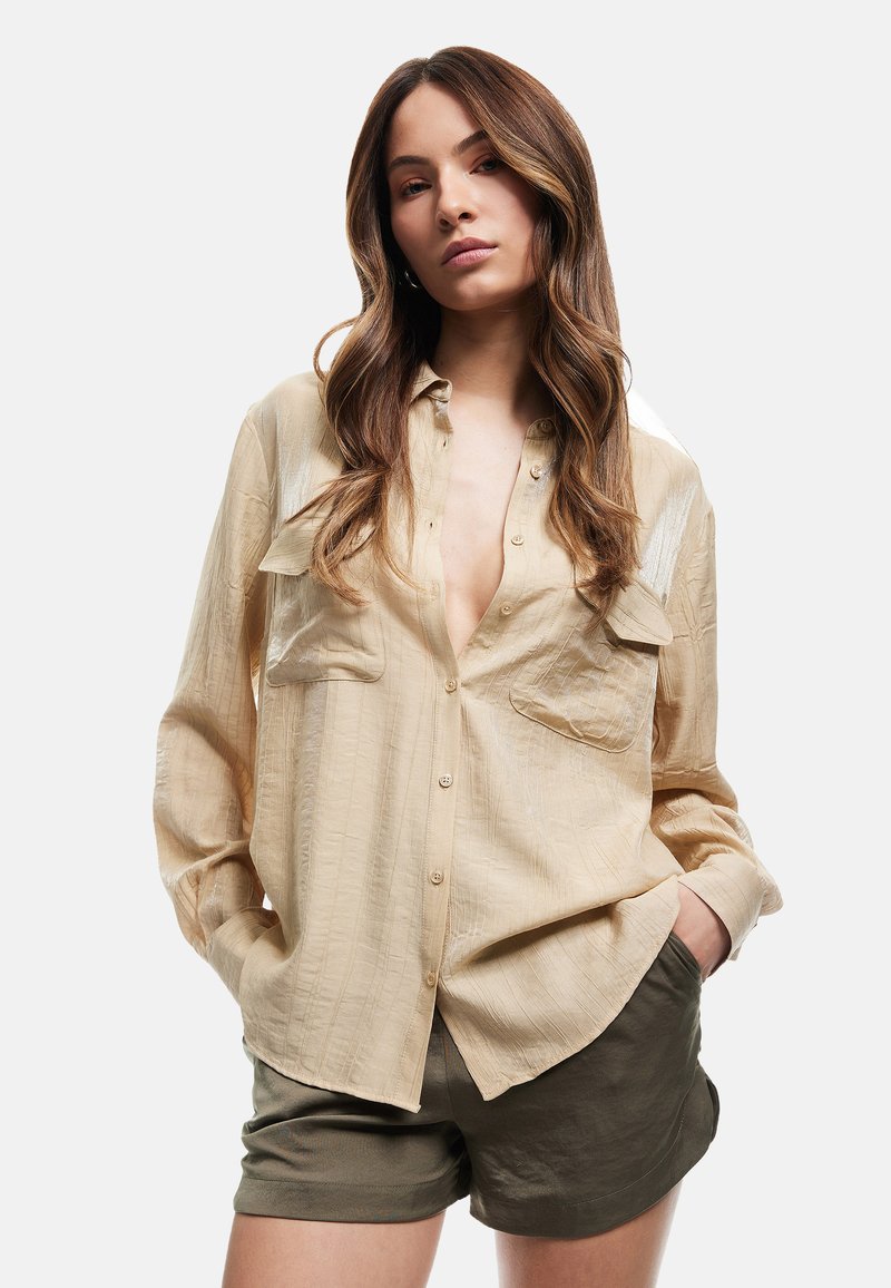 Ljus beige långärmad skjorta med två bröstfickor, knäppning framtill, tillverkad av texturerat tyg. I kombination med mörkgröna shorts.