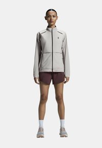 Chaqueta deportiva gris claro con cremallera frontal, detalles en negro contrastantes y un diseño ajustado, combinada con pantalones cortos oscuros y zapatillas deportivas.