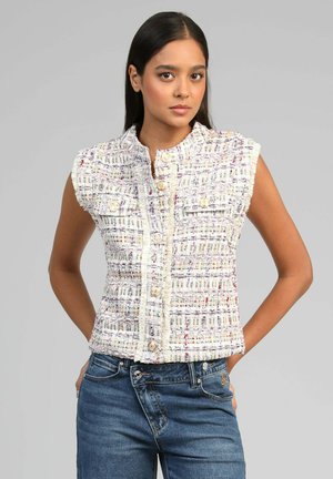 Jeune femme aux longs cheveux noirs portant un gilet sans manches en tweed multicolore à boutons et un jean bleu denim, sur un fond gris uni.